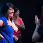 Two women stand, smiling, with their hands on their hearts as they listen to a storyteller.